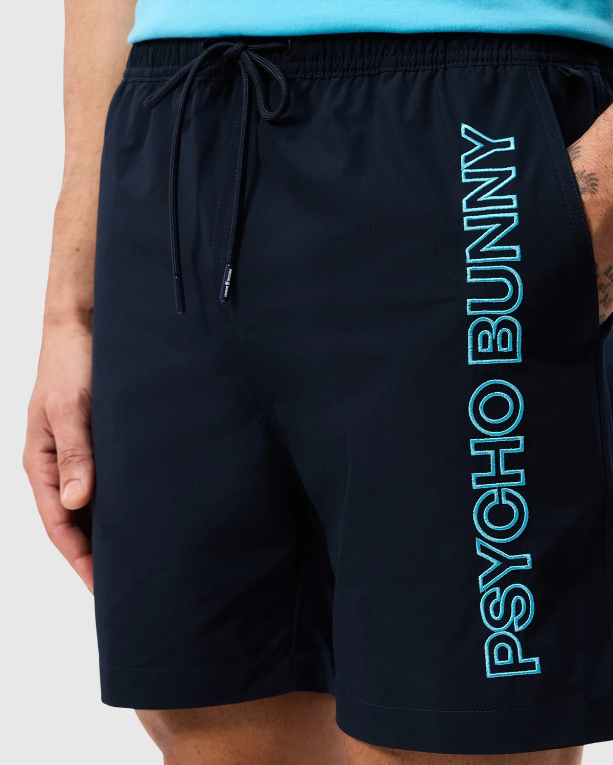 Close-up shot of a person wearing Psycho Bunny MENS MELVIN SWIM TRUNK - B6W665C200 in black, featuring the words "PSYCHO BUNNY" vertically printed in light blue on the right leg. The individual has their right hand in their pocket and is paired with a turquoise top. The quick-dry outer shell and drawstring stretch waistband offer both style and comfort.