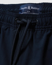 Close-up shot of navy blue Psycho Bunny Melvin Swim Trunks (product code: B6W665C200) featuring a stretch waistband and drawstring. The label at the waistband reads "Psycho Bunny" with a small bunny logo and indicates the size as "M-M." The label also mentions the quick-dry outer shell and that the product is made in China.
