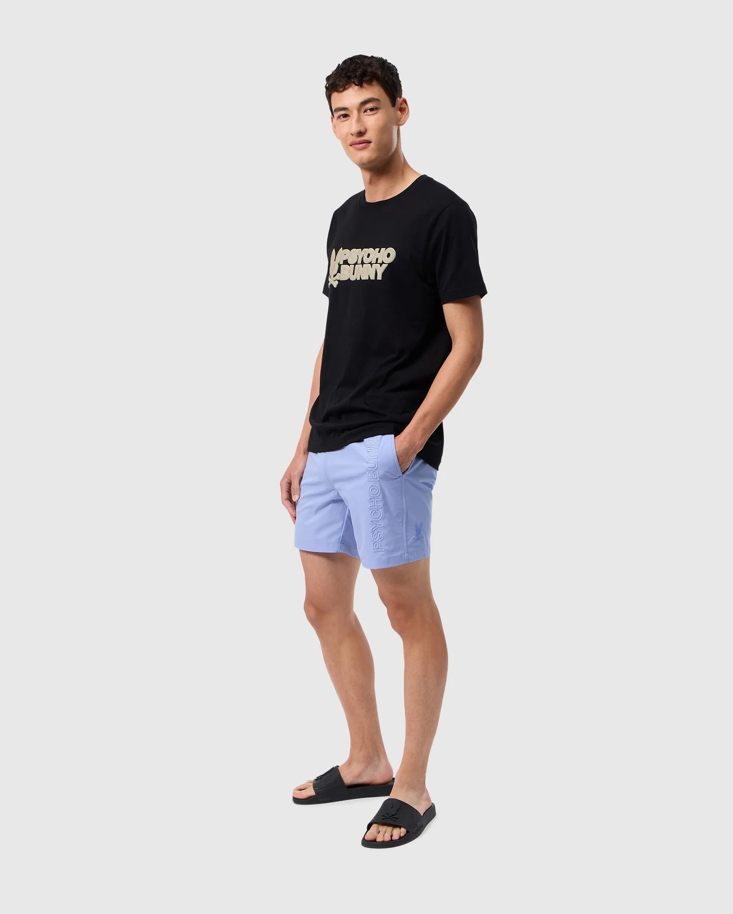 A young man stands against a plain, light gray background. He is wearing a black graphic T-shirt with text, the Psycho Bunny MENS MELVIN SWIM TRUNK - B6W665C200 in light blue, featuring pockets and made of quick-dry textile, and black slide sandals. He has short, dark hair and is smiling slightly while looking at the camera.
