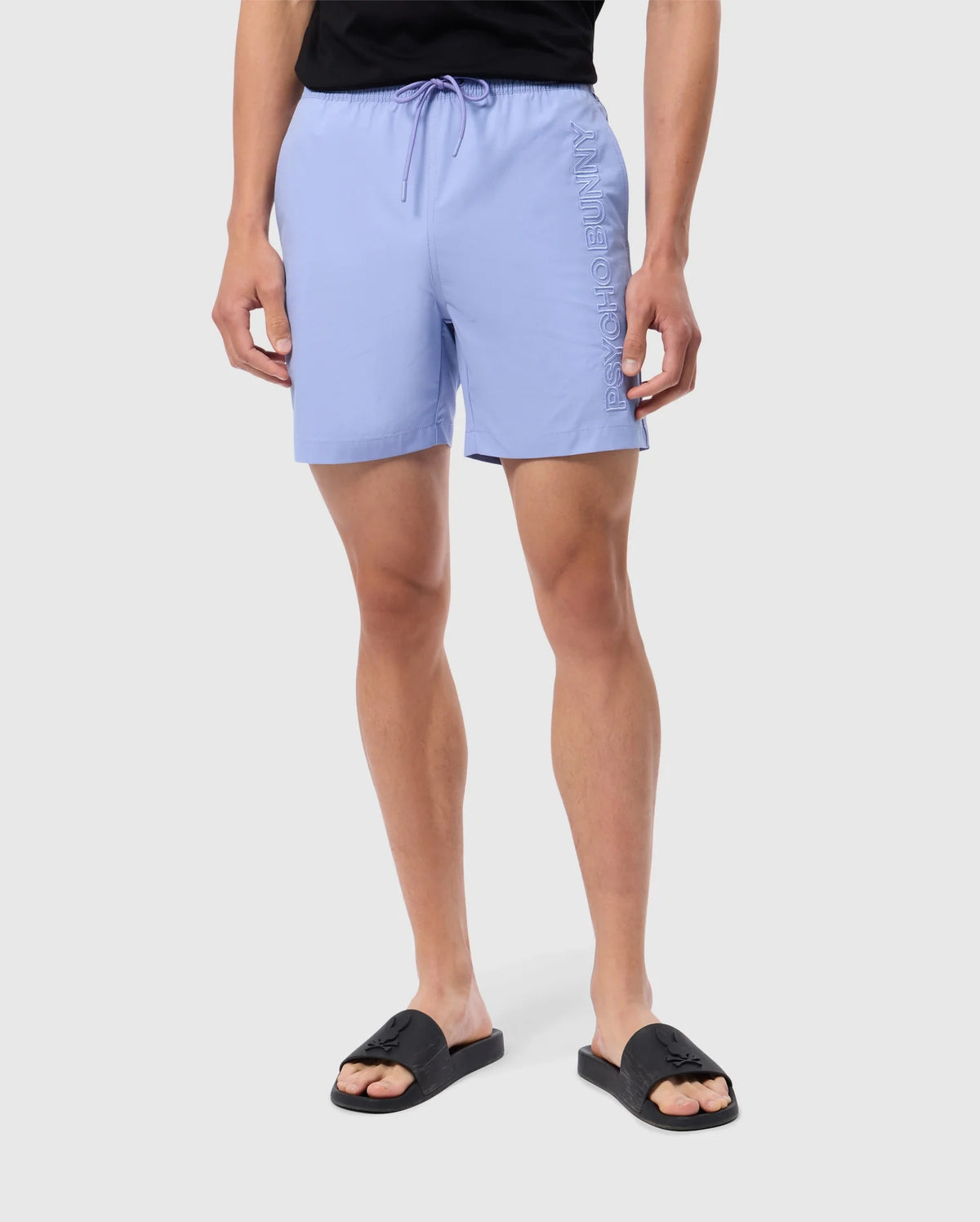 A person is standing against a plain background, wearing light blue MENS MELVIN SWIM TRUNK - B6W665C200 by Psycho Bunny with the brand name embroidered along the side. They are also wearing a black t-shirt and black slide sandals, while holding a waterproof bag. Their hands are relaxed by their sides.