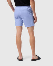 A person stands facing away from the camera, wearing a black t-shirt, black flip-flops, and Psycho Bunny's light blue MENS MELVIN SWIM TRUNK - B6W665C200 made of quick-dry textile. The shorts feature an elastic waistband and a single back pocket on the right side. The background is plain white.