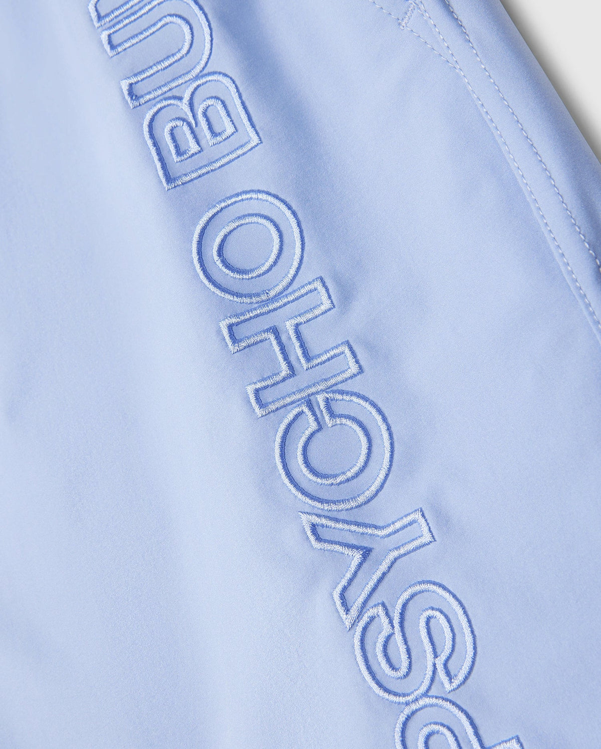 Close-up of light blue fabric from the Psycho Bunny MENS MELVIN SWIM TRUNK - B6W665C200, featuring the embroidered text "PSYCHO BUN" in white, partially capturing the word "PSYCHO BUNNY" along a seam. The stitching and material texture of this quick-dry textile are clearly visible.