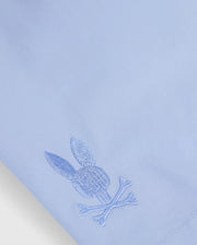 Close-up of a light blue quick-dry textile from the MENS MELVIN SWIM TRUNK - B6W665C200 by Psycho Bunny, featuring an embroidered logo of a rabbit head with ears above two crossed bones, near the bottom corner. The embroidery is done in a matching blue thread.