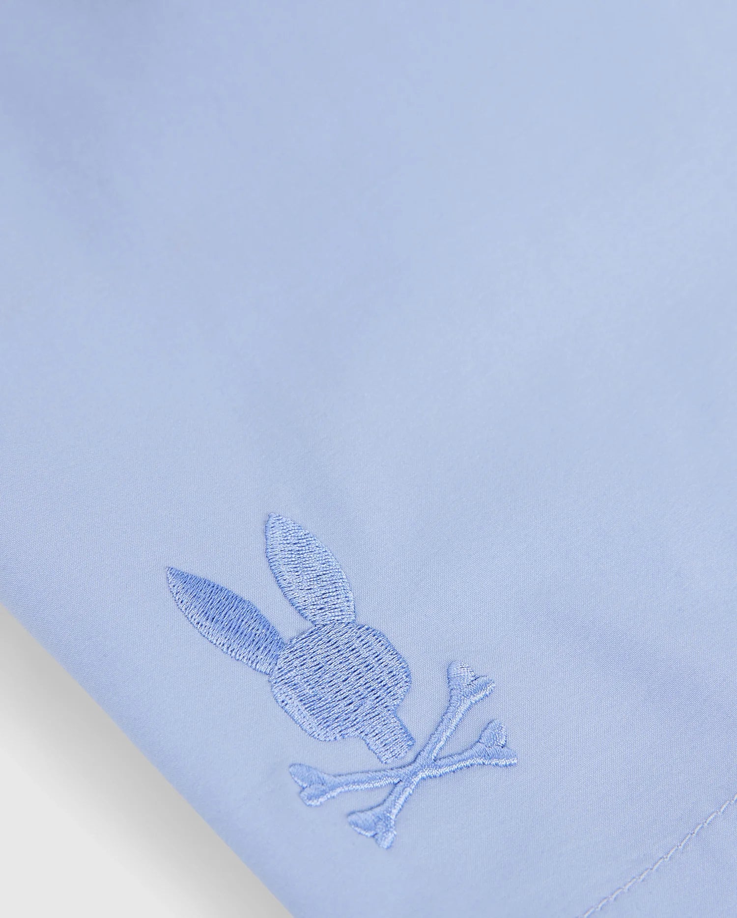 Close-up of a light blue quick-dry textile from the MENS MELVIN SWIM TRUNK - B6W665C200 by Psycho Bunny, featuring an embroidered logo of a rabbit head with ears above two crossed bones, near the bottom corner. The embroidery is done in a matching blue thread.