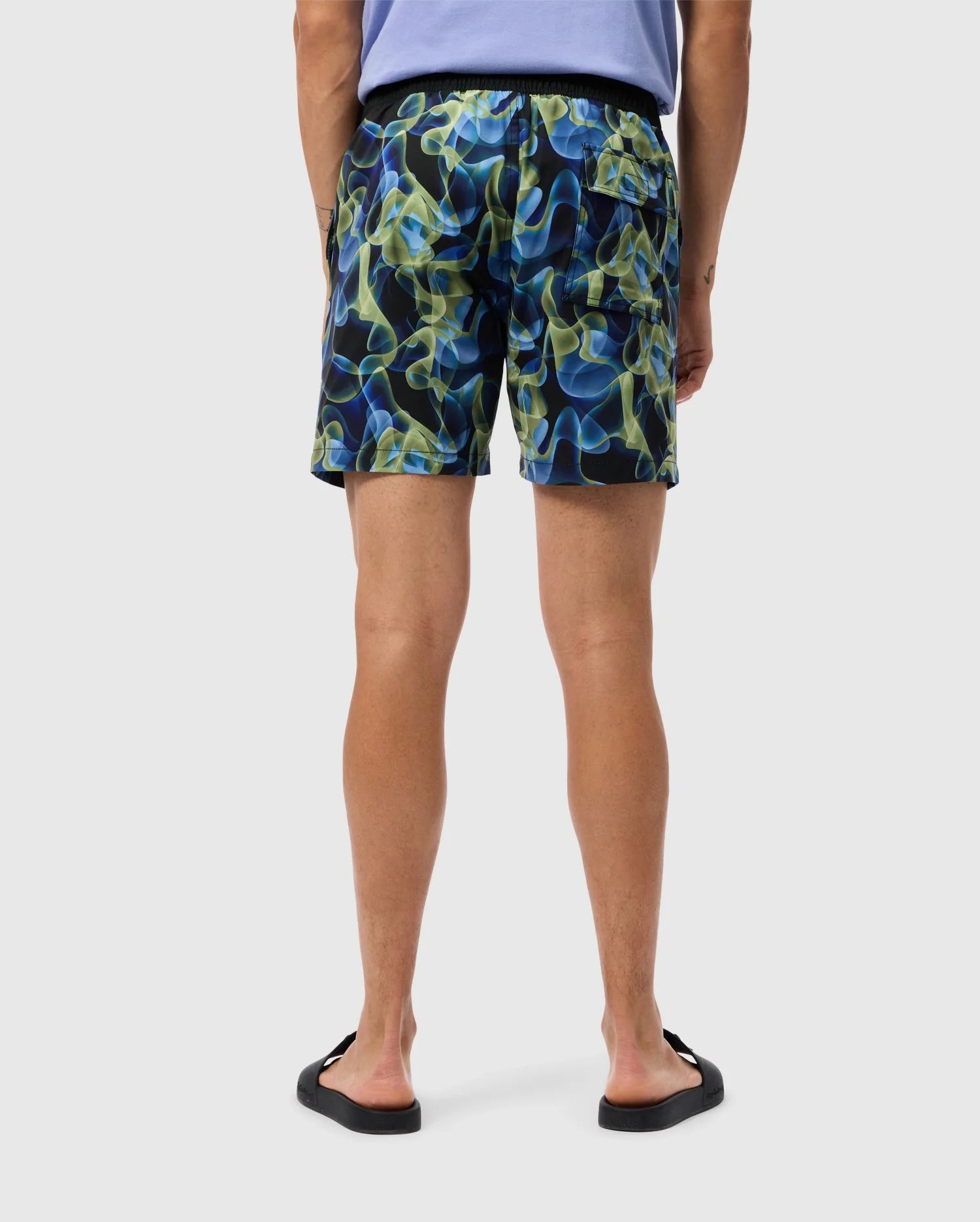 A person is standing and facing away, wearing a light purple T-shirt, Psycho Bunny's MENS GOLDCREEK ALL OVER PRINT SWIM TRUNK - B6W672C200 in blue, green, and yellow abstract patterns, and black flip-flops. The background is plain and light-colored.