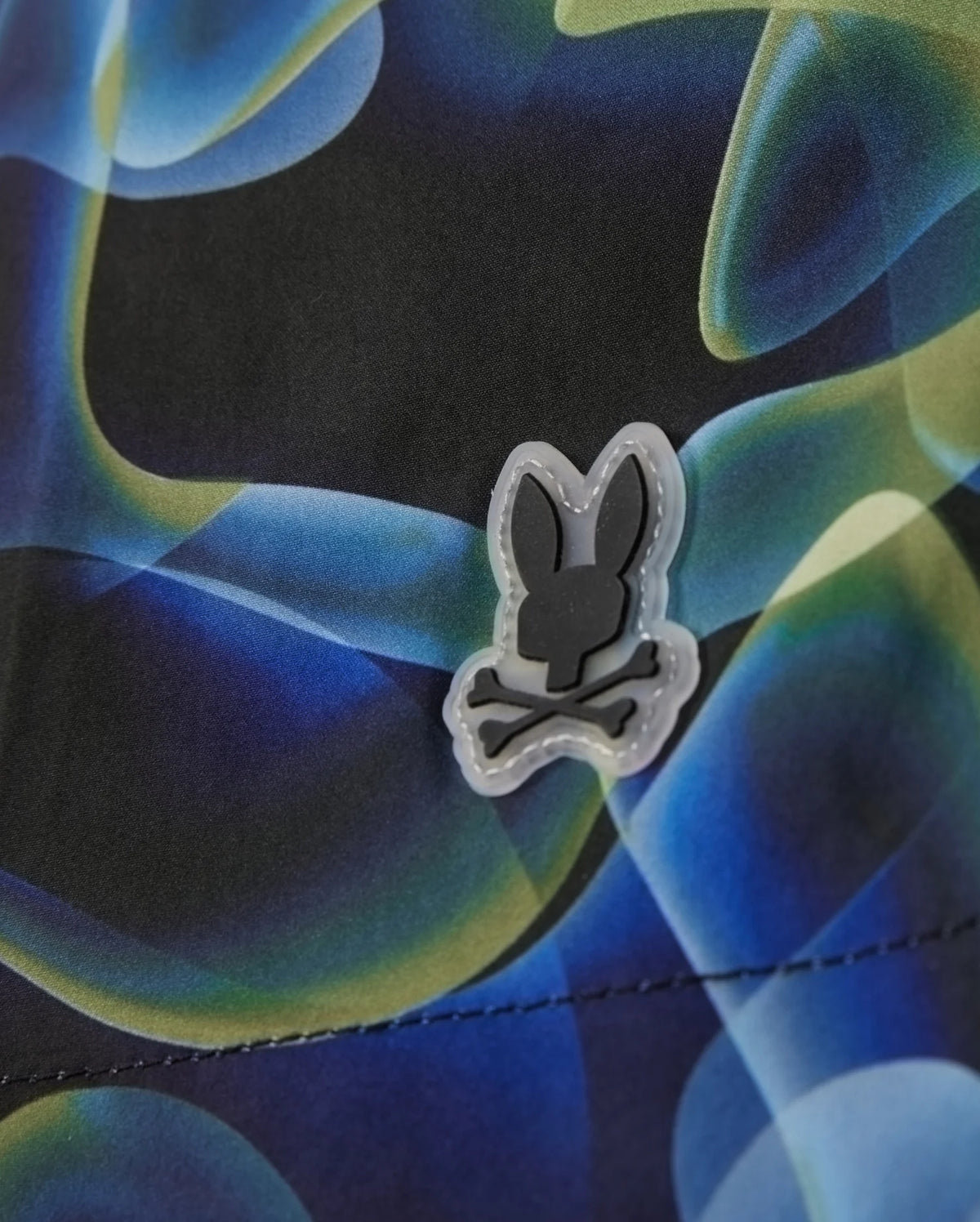 These Psycho Bunny MENS GOLDCREEK ALL OVER PRINT SWIM TRUNK - B6W672C200 feature a colorful abstract design with blue, green, and black hues. The fabric is adorned with a small patch showcasing a bunny head with crossbones below it. The stitching of this super-comfy swimwear is visible at the bottom of this unique image.