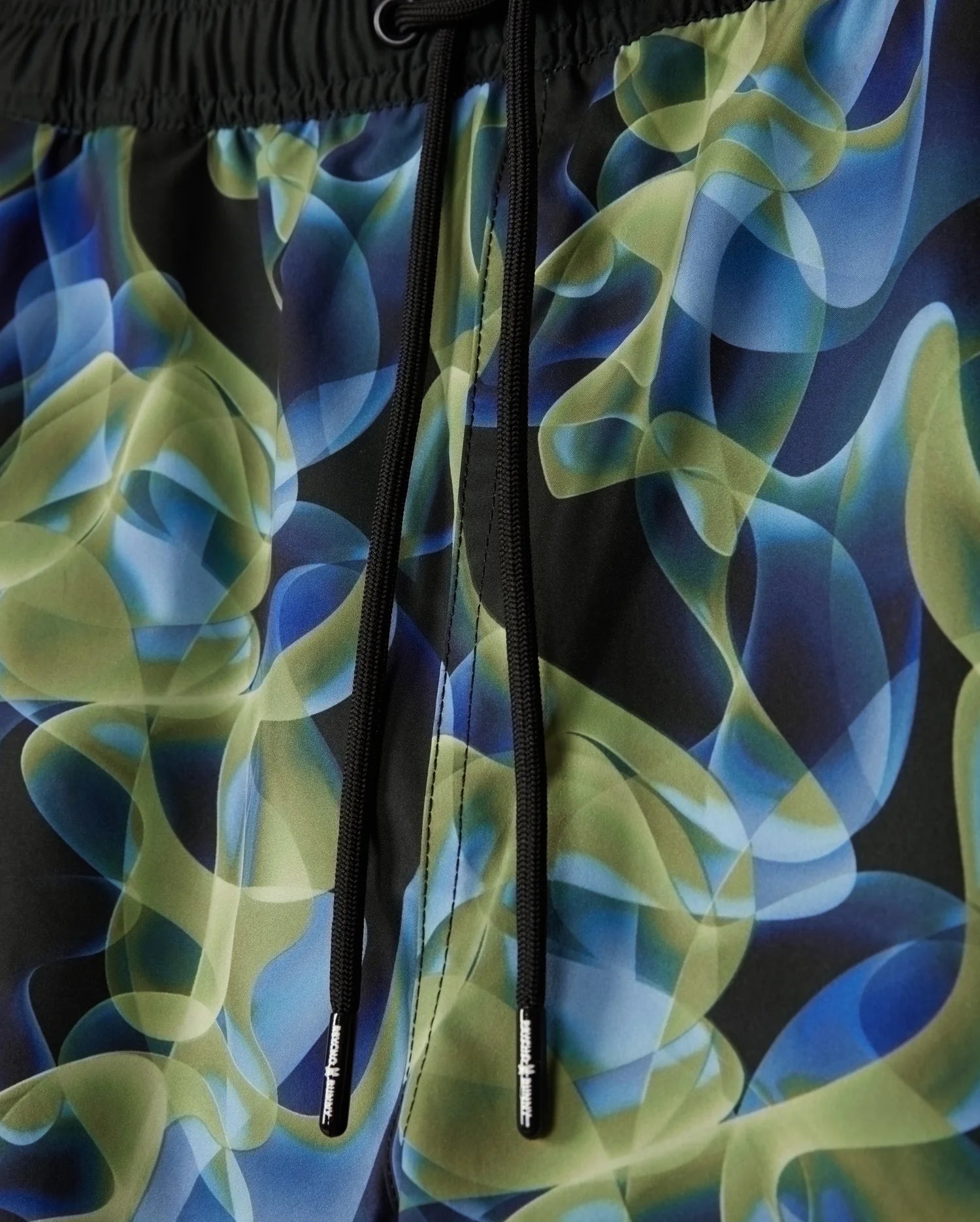Detailed look at the MENS GOLDCREEK ALL OVER PRINT SWIM TRUNK - B6W672C200 by Psycho Bunny, showcasing a vibrant abstract pattern with swirling shapes in shades of green, blue, and black. The swim trunks feature black drawstrings with small metallic end caps for added style.