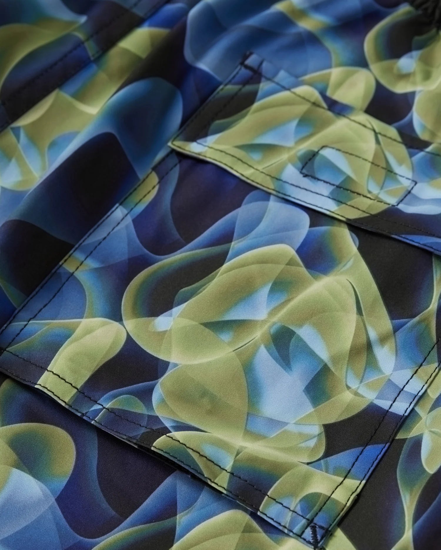 Close-up of a Psycho Bunny fabric featuring an abstract pattern with swirling green and blue shapes, reminiscent of the MENS GOLDCREEK ALL OVER PRINT SWIM TRUNK - B6W672C200. This super-comfy swim piece includes a pocket with visible stitching. The colors blend smoothly, creating a vibrant and fluid design.