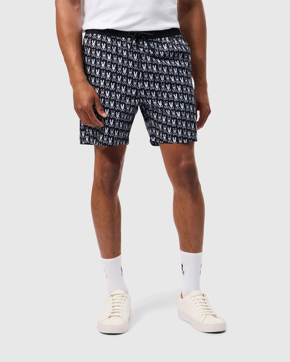 A person is shown wearing Psycho Bunny's MENS TILDEN PRINT SWIM TRUNK (B6W729D200) featuring a repeating white pattern, white socks, and white sneakers. Their upper body is not visible. The contrast bunny print adds a playful touch against the plain, light gray background.