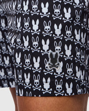 Close-up of a person wearing Psycho Bunny MENS TILDEN PRINT SWIM TRUNKS - B6W729D200, adorned with a white skull and crossbones pattern that features bunny ears on the skulls. A small, reflective black bunny-shaped logo is also visible on the bottom hem of these contrast bunny print shorts. The person's leg is partially shown.