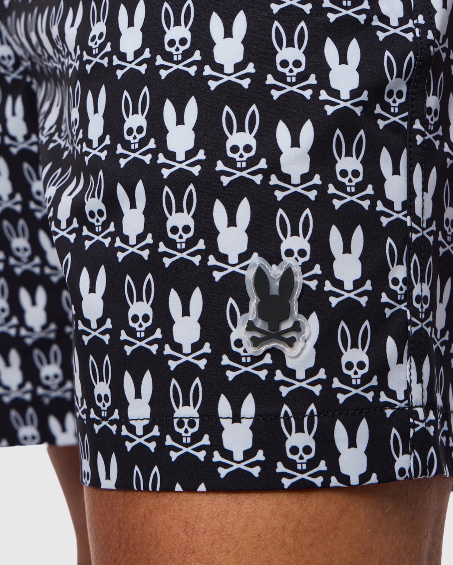 Close-up of a person wearing Psycho Bunny MENS TILDEN PRINT SWIM TRUNKS - B6W729D200, adorned with a white skull and crossbones pattern that features bunny ears on the skulls. A small, reflective black bunny-shaped logo is also visible on the bottom hem of these contrast bunny print shorts. The person's leg is partially shown.