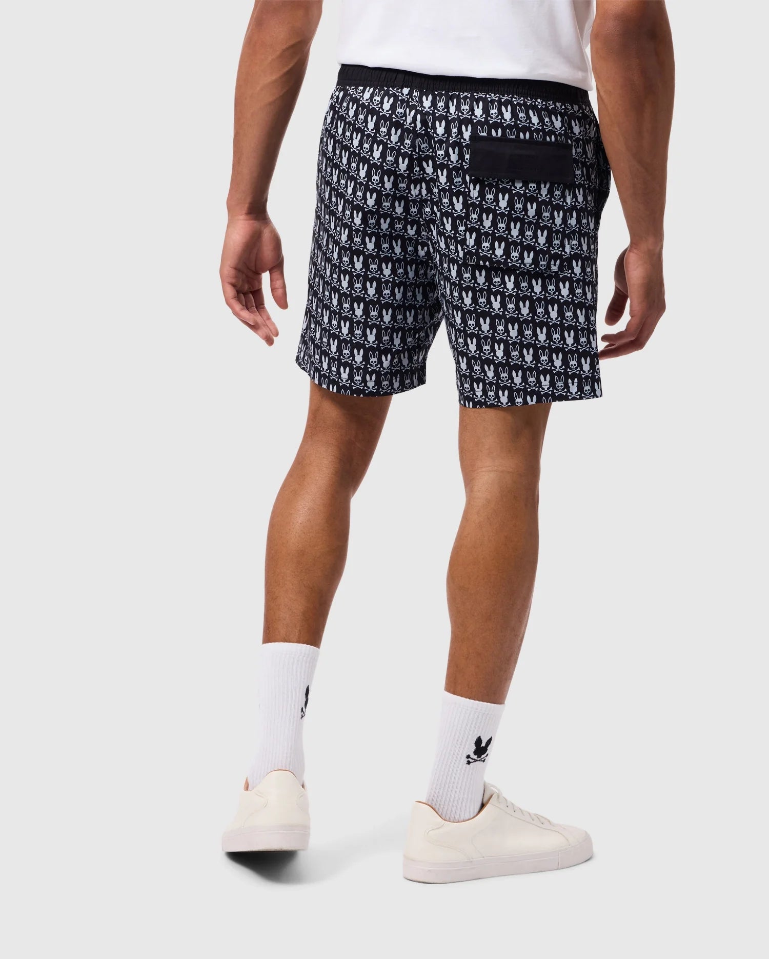 A person stands with their back to the camera, sporting a white T-shirt and black-and-white Psycho Bunny Men's Tilden Print Swim Trunks (Product Code: B6W729D200) featuring a contrast bunny print. They pair this look with white socks and sneakers. The swim trunks boast a repeating logo design and include a back pocket.