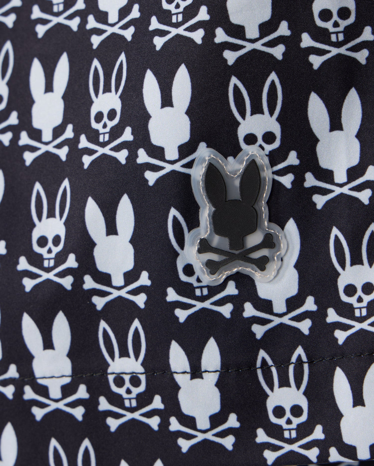 A close-up view of the MENS TILDEN PRINT SWIM TRUNK by Psycho Bunny (B6W729D200) reveals a fabric pattern of white skulls with bunny ears and crossbones, set against a dark background. A rubberized patch with a similar design is sewn onto the material. The contrast bunny print repeats uniformly across the quick-dry membrane, making these shorts perfect for any swim.