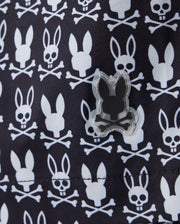 A close-up view of the MENS TILDEN PRINT SWIM TRUNK by Psycho Bunny (B6W729D200) reveals a fabric pattern of white skulls with bunny ears and crossbones, set against a dark background. A rubberized patch with a similar design is sewn onto the material. The contrast bunny print repeats uniformly across the quick-dry membrane, making these shorts perfect for any swim.