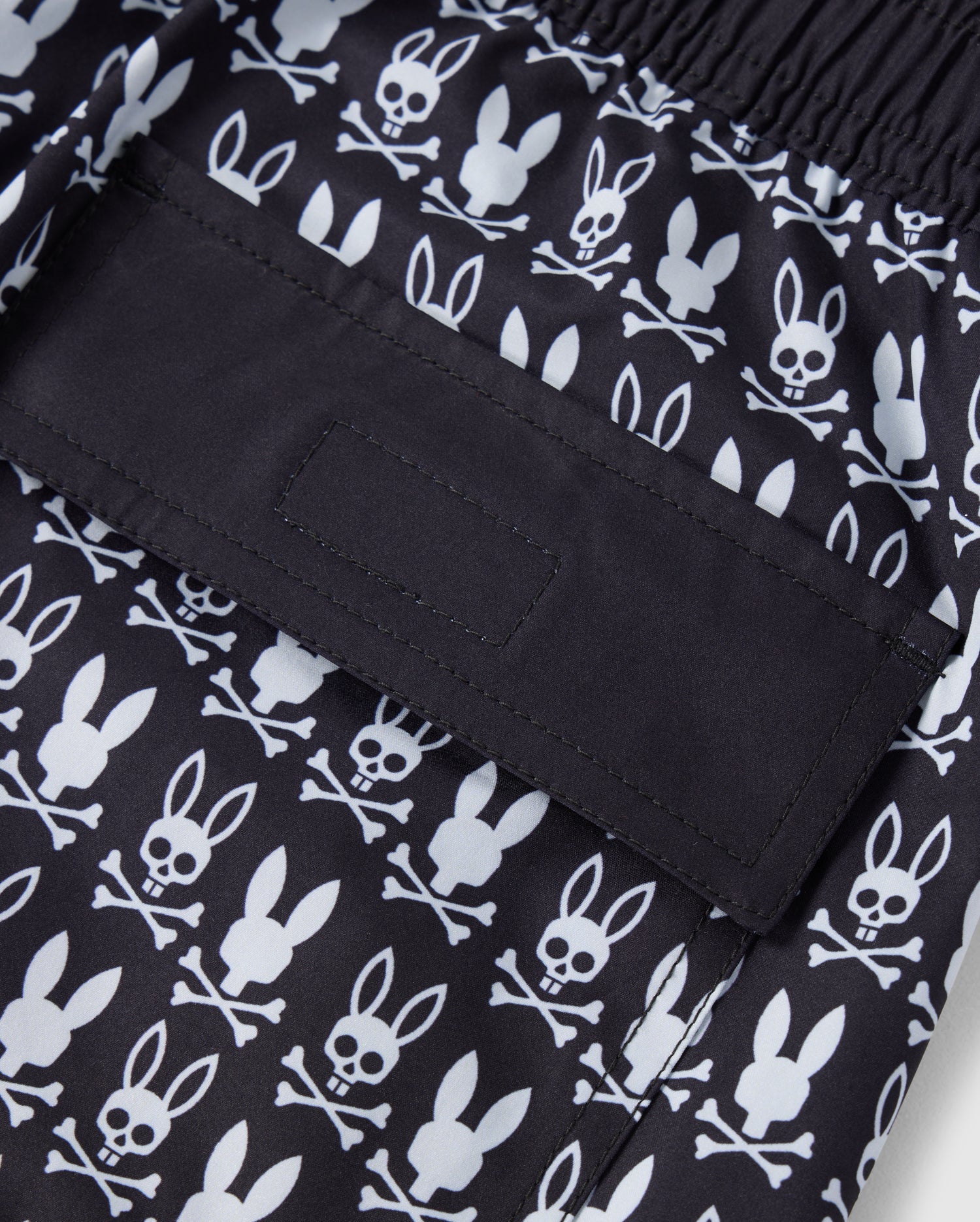 A close-up of the Psycho Bunny MENS TILDEN PRINT SWIM TRUNK - B6W729D200 featuring a contrast bunny print with white cartoon-style bunny skulls and crossbones. The image focuses on a back pocket with a flap secured by Velcro. The elastic waistband makes these swim trunks perfect for any casual or beach setting.