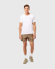 A man stands against a neutral background. He is wearing a plain white t-shirt and green Psycho Bunny "MENS ARNOLD PRINT SWIM TRUNK - B6W820D200" featuring an orange circular pattern. He has short, styled hair and is wearing tan sneakers with white socks. The trunks, made from quick-dry material, feature a comfortable stretch mesh lining. He is looking at the camera with a neutral expression.
