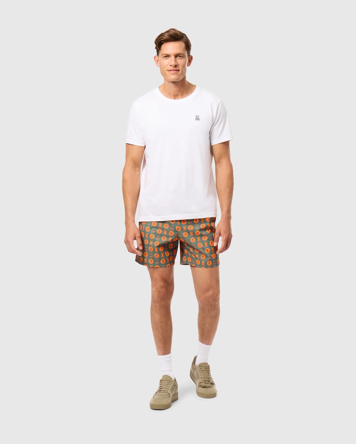 A man stands against a neutral background. He is wearing a plain white t-shirt and green Psycho Bunny "MENS ARNOLD PRINT SWIM TRUNK - B6W820D200" featuring an orange circular pattern. He has short, styled hair and is wearing tan sneakers with white socks. The trunks, made from quick-dry material, feature a comfortable stretch mesh lining. He is looking at the camera with a neutral expression.
