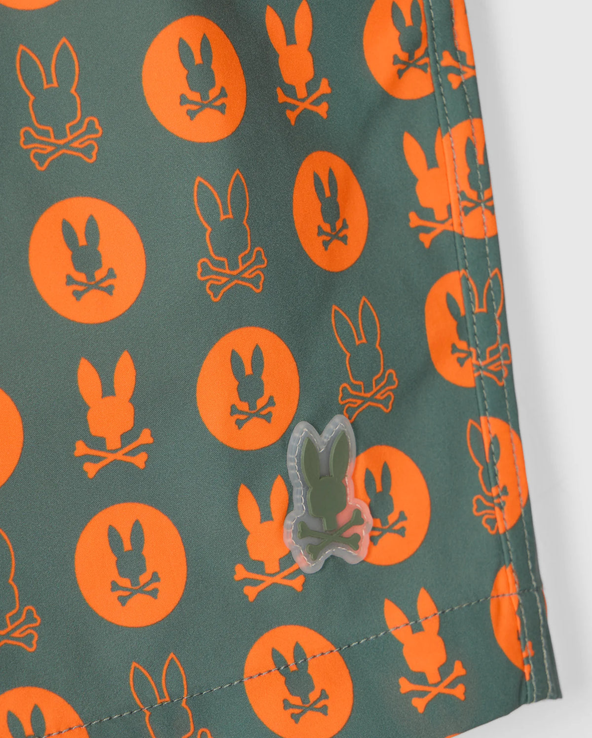 Close-up of the Psycho Bunny MENS ARNOLD PRINT SWIM TRUNK - B6W820D200, featuring a green base with an orange pattern of bunny skulls and crossbones inside circles. The quick-dry material boasts a transparent rubberized logo sewn onto the corner. A comfortable stretch mesh lining adds to the overall ease and functionality.