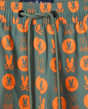 Close-up of Psycho Bunny's MENS ARNOLD PRINT SWIM TRUNK (B6W820D200), showcasing green swim trunks adorned with an orange pattern featuring a cartoon rabbit head and crossbones inside circles. A green drawstring is tied at the center of an elastic waistband, and they're crafted from quick-dry material for your convenience.