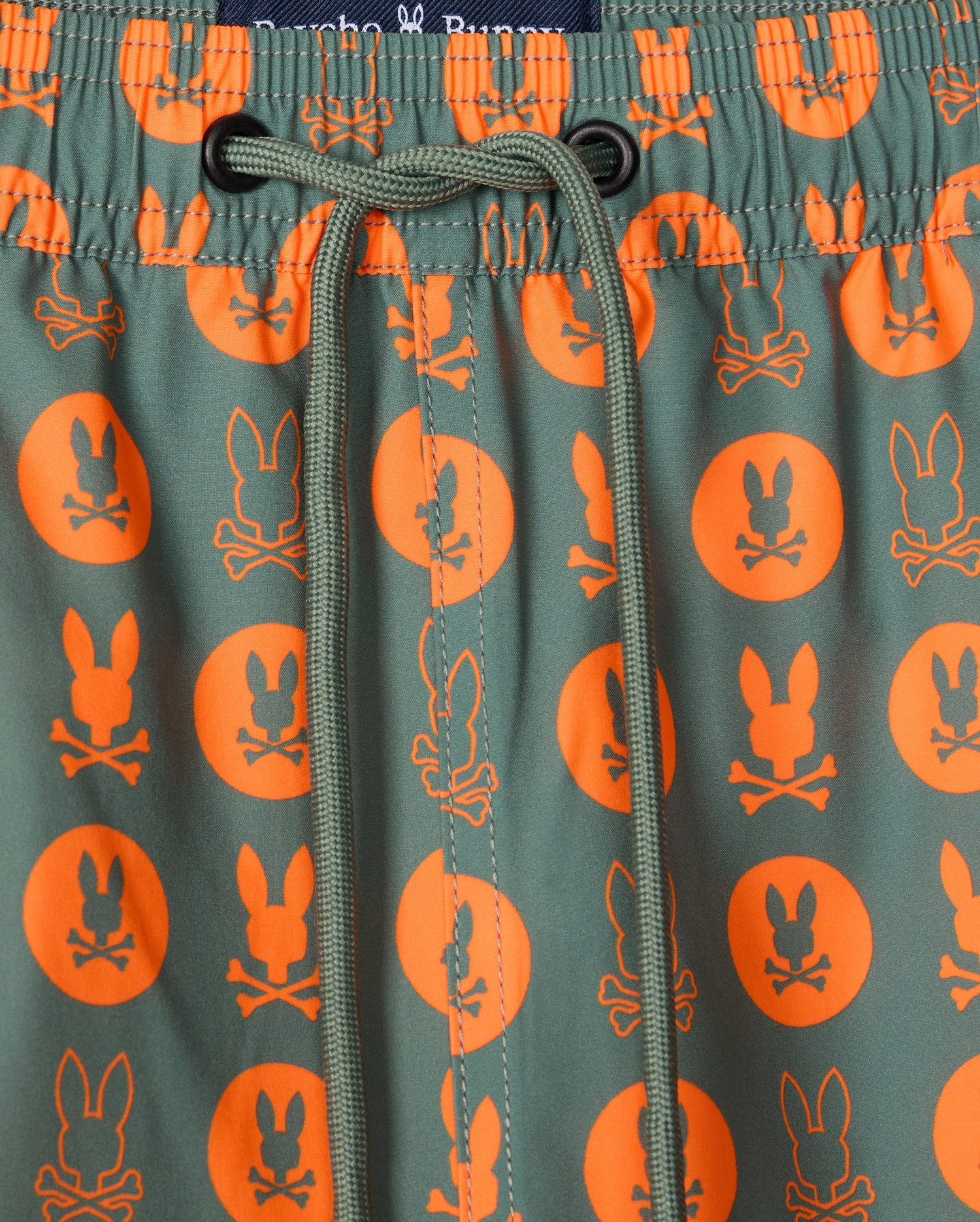 Close-up of Psycho Bunny's MENS ARNOLD PRINT SWIM TRUNK (B6W820D200), showcasing green swim trunks adorned with an orange pattern featuring a cartoon rabbit head and crossbones inside circles. A green drawstring is tied at the center of an elastic waistband, and they're crafted from quick-dry material for your convenience.