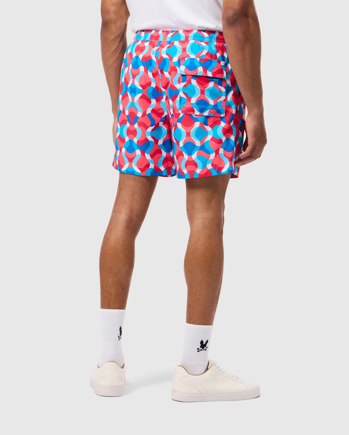 A person is standing with their back to the camera, wearing a white t-shirt and vibrant MENS WALTER LIGHTWEIGHT PRINT SWIM TRUNK - B6W863D200 by Psycho Bunny in blue and red. They complete the look with white socks and sneakers. The background is plain white.