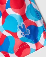 Close-up of a fabric showcasing a dynamic pattern of overlapping circles in red, blue, white, and light blue. A small blue rabbit skull and crossbones emblem is stitched onto the 100% polyester material near a seam, echoing the playful style of the Psycho Bunny MENS WALTER LIGHTWEIGHT PRINT SWIM TRUNK - B6W863D200.