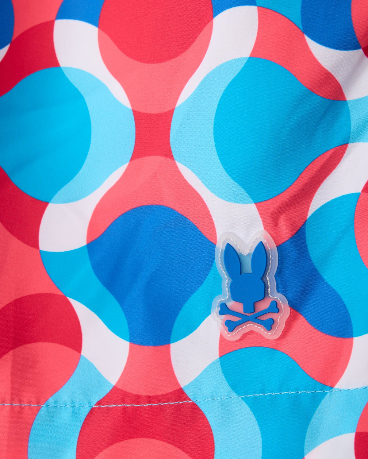 The MENS WALTER LIGHTWEIGHT PRINT SWIM TRUNK - B6W863D200 from Psycho Bunny showcases a vibrant design with large, overlapping blue, white, and red circles. Crafted from 100% polyester, it features a unique detail in the lower right center: a small patch with a blue bunny skull and crossbones symbol.
