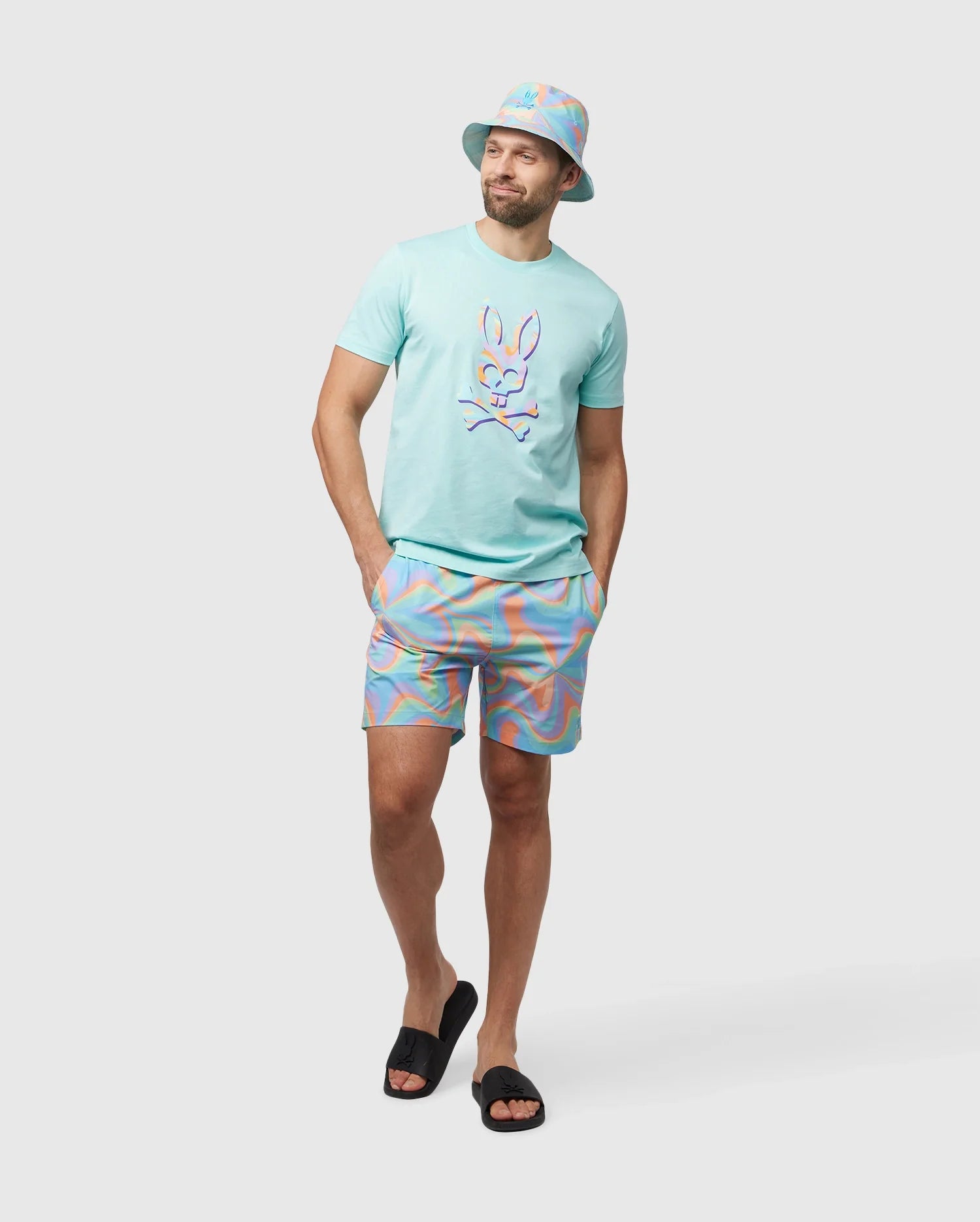 A man is wearing a pastel-colored outfit, including a bucket hat, a t-shirt with a cartoon rabbit graphic, and quick-drying Psycho Bunny MENS BLOOMINGTON SWIM TRUNK - B6W941A2PO with a swirl pattern. He is standing against a light gray background with his hands in his pockets and wearing black slides.