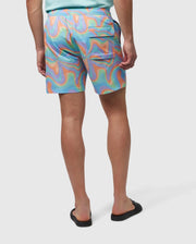 A person is seen from behind, wearing colorful, swirly-patterned Psycho Bunny MENS BLOOMINGTON SWIM TRUNK - B6W941A2PO in shades of blue, orange, and green. They also wear a light teal shirt and black slip-on shoes. A small silicone bunny patch adds a quirky touch. The background is plain white.