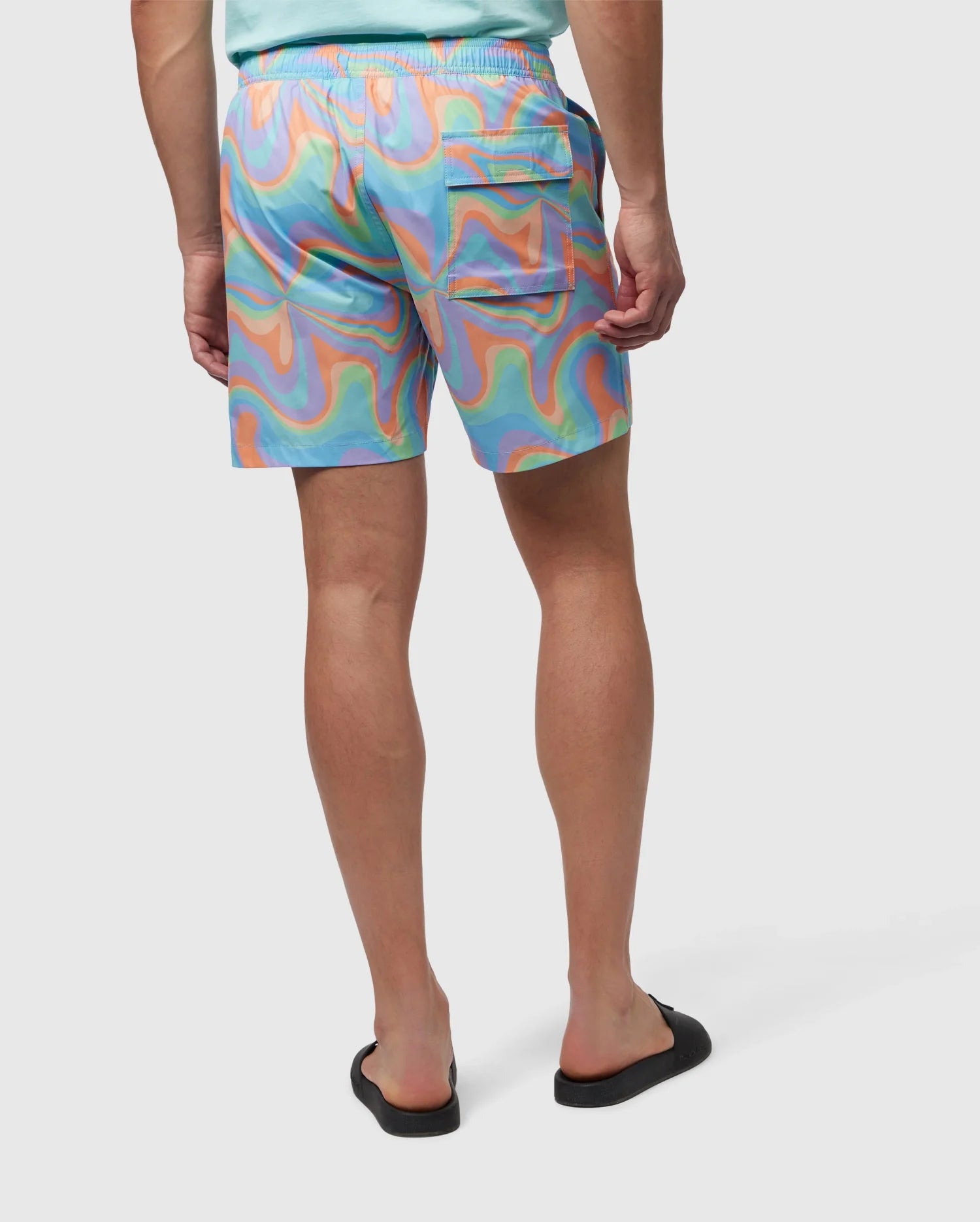 A person is seen from behind, wearing colorful, swirly-patterned Psycho Bunny MENS BLOOMINGTON SWIM TRUNK - B6W941A2PO in shades of blue, orange, and green. They also wear a light teal shirt and black slip-on shoes. A small silicone bunny patch adds a quirky touch. The background is plain white.
