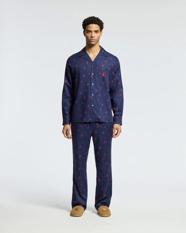 A man stands wearing a matching Men's All Over Print Flannel Pajama Set in navy blue with a red pattern. The soft cotton flannel sleepwear by Psycho Bunny features a button-up top and full-length pants, complemented perfectly by his brown slippers. The background is plain white.