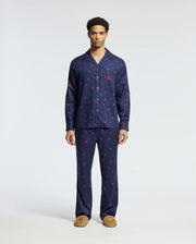 A man stands wearing a matching Men's All Over Print Flannel Pajama Set in navy blue with a red pattern. The soft cotton flannel sleepwear by Psycho Bunny features a button-up top and full-length pants, complemented perfectly by his brown slippers. The background is plain white.
