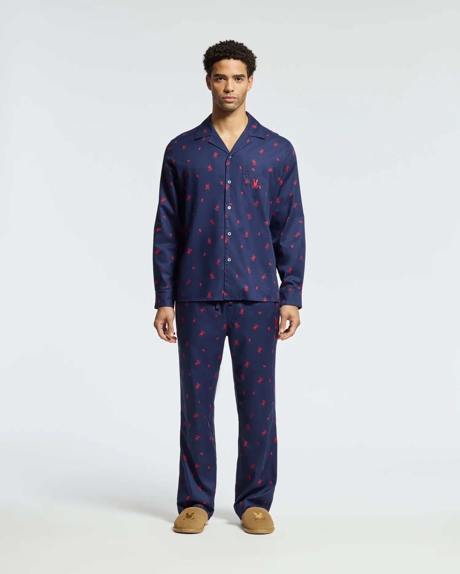 A man stands wearing a matching Men's All Over Print Flannel Pajama Set in navy blue with a red pattern. The soft cotton flannel sleepwear by Psycho Bunny features a button-up top and full-length pants, complemented perfectly by his brown slippers. The background is plain white.
