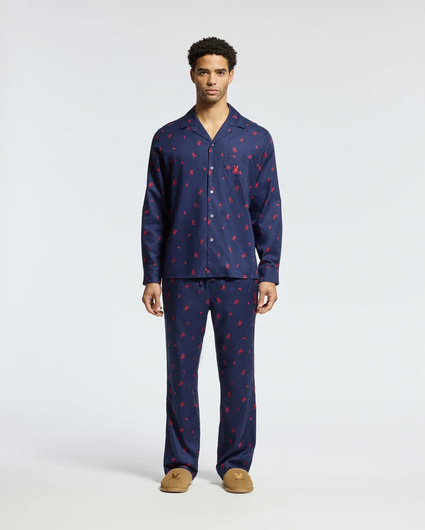 A man stands wearing a matching Men's All Over Print Flannel Pajama Set in navy blue with a red pattern. The soft cotton flannel sleepwear by Psycho Bunny features a button-up top and full-length pants, complemented perfectly by his brown slippers. The background is plain white.