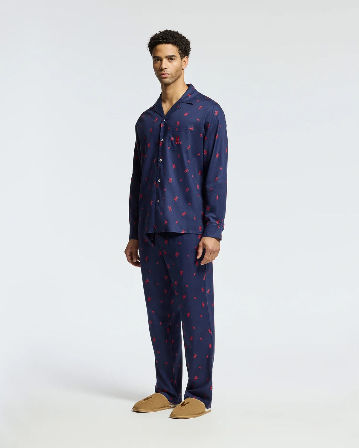 A man wearing a navy Psycho Bunny Men's All Over Print Flannel Pajama Set (B6X343E200) with small red patterns stands against a plain background. He pairs this soft flannel sleep set with tan slippers.