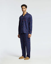 A man wearing a navy Psycho Bunny Men's All Over Print Flannel Pajama Set (B6X343E200) with small red patterns stands against a plain background. He pairs this soft flannel sleep set with tan slippers.