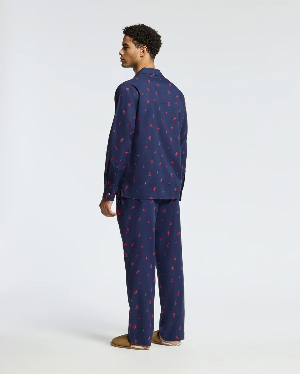 A man stands facing sideways, wearing the Psycho Bunny MENS ALL OVER PRINT FLANNEL PAJAMA SET (B6X343E200) in dark blue adorned with a subtle red pattern. The soft flannel sleep set complements his brown slippers against the plain, light-colored background.