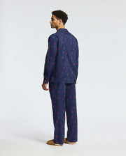 A man stands facing sideways, wearing the Psycho Bunny MENS ALL OVER PRINT FLANNEL PAJAMA SET (B6X343E200) in dark blue adorned with a subtle red pattern. The soft flannel sleep set complements his brown slippers against the plain, light-colored background.