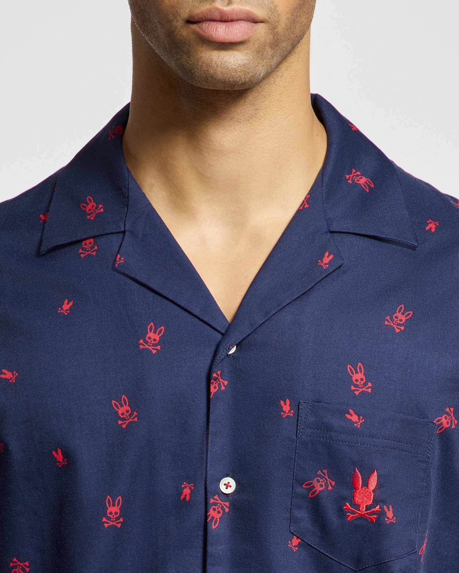 The individual is adorned in a shirt from the Psycho Bunny Men's All Over Print Flannel Pajama Set, B6X343E200, characterized by its navy color adorned with red embroidered bunny motifs. The shirt includes a collar, button closure, and chest pocket, blending stylish elements with the comforting feel of cotton flannel sleepwear.