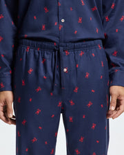 A person is wearing the Psycho Bunny MENS ALL OVER PRINT FLANNEL PAJAMA SET - B6X343E200, featuring soft cotton flannel with a design of small red bunny patterns. This sleepwear set includes a long-sleeve button-up top and pants equipped with an elastic waistband and drawstring, emphasizing the midsection and partially revealing the arms and hands.