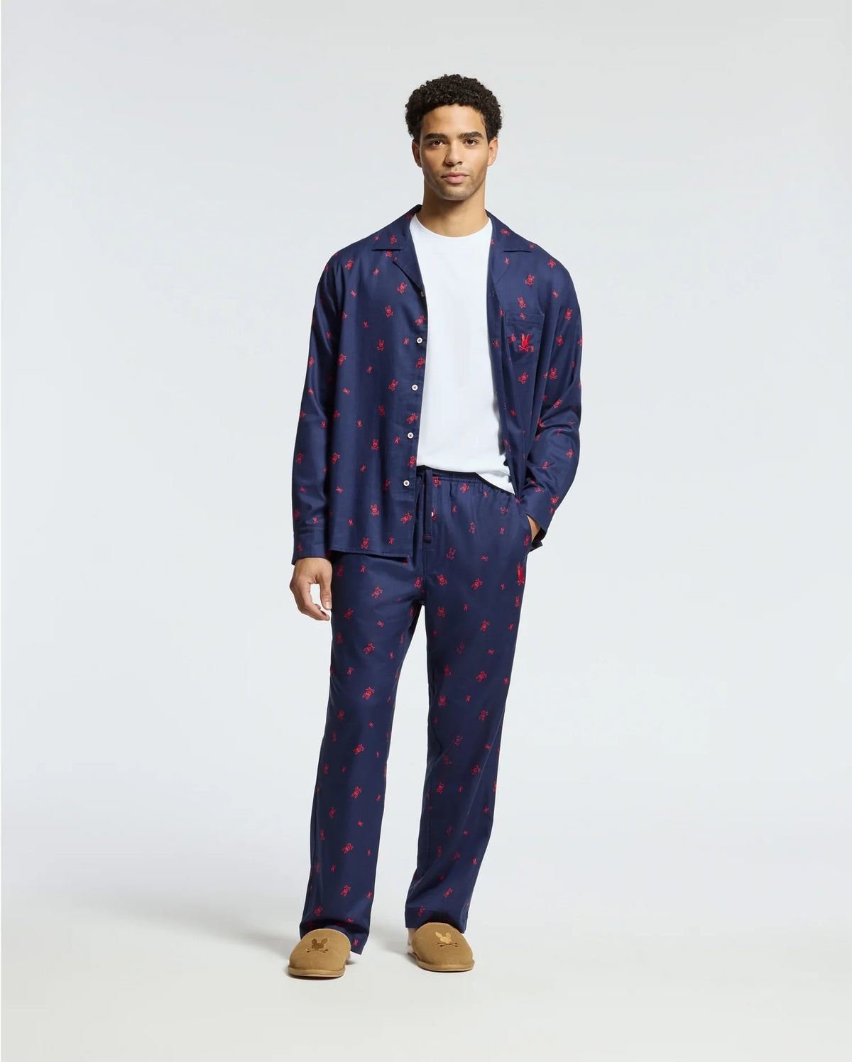 A man is standing in a plain white background, dressed in a navy blue Psycho Bunny MENS ALL OVER PRINT FLANNEL PAJAMA SET - B6X343E200 featuring red prints, complemented by a white t-shirt and brown slippers.