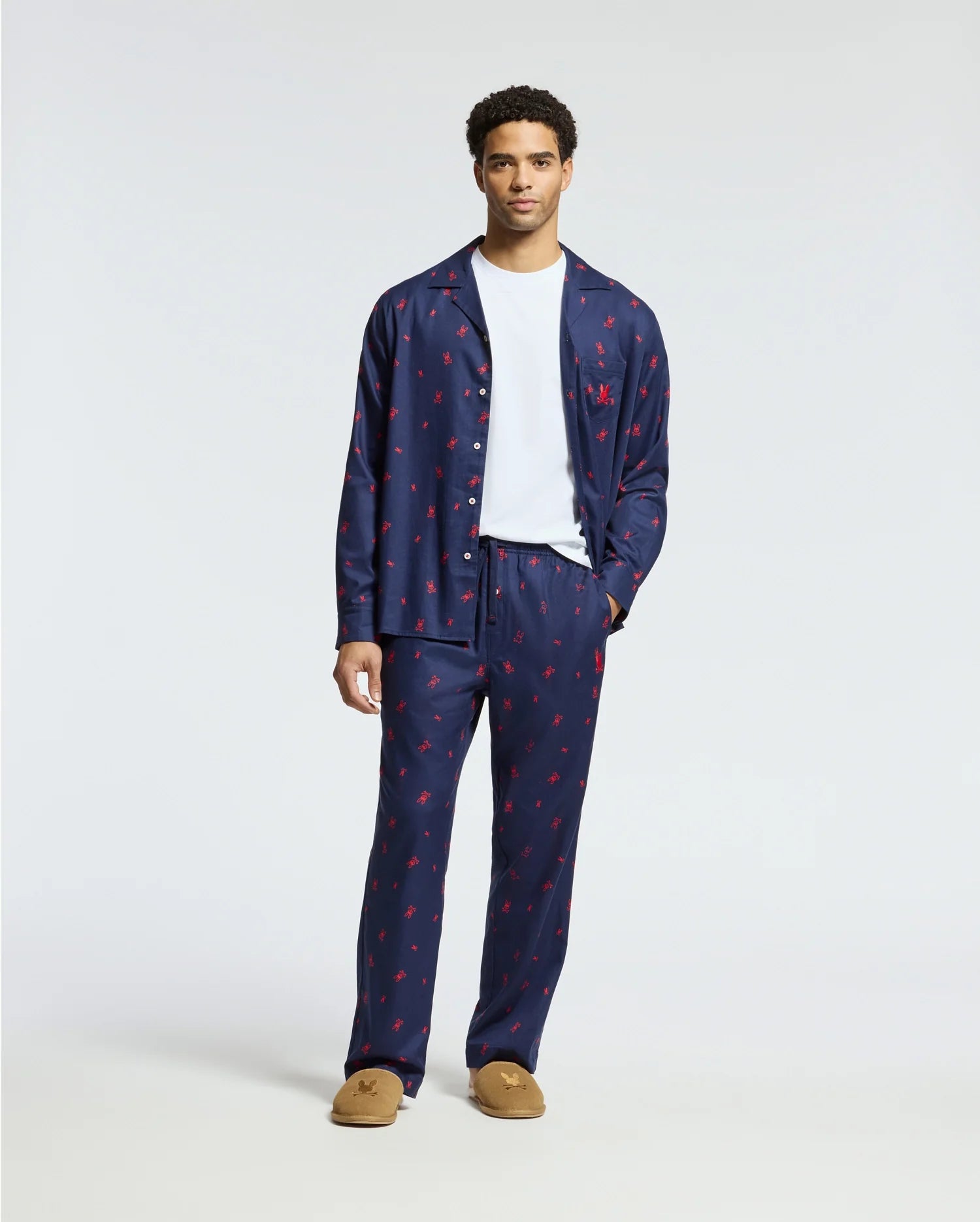 A man is standing in a plain white background, dressed in a navy blue Psycho Bunny MENS ALL OVER PRINT FLANNEL PAJAMA SET - B6X343E200 featuring red prints, complemented by a white t-shirt and brown slippers.