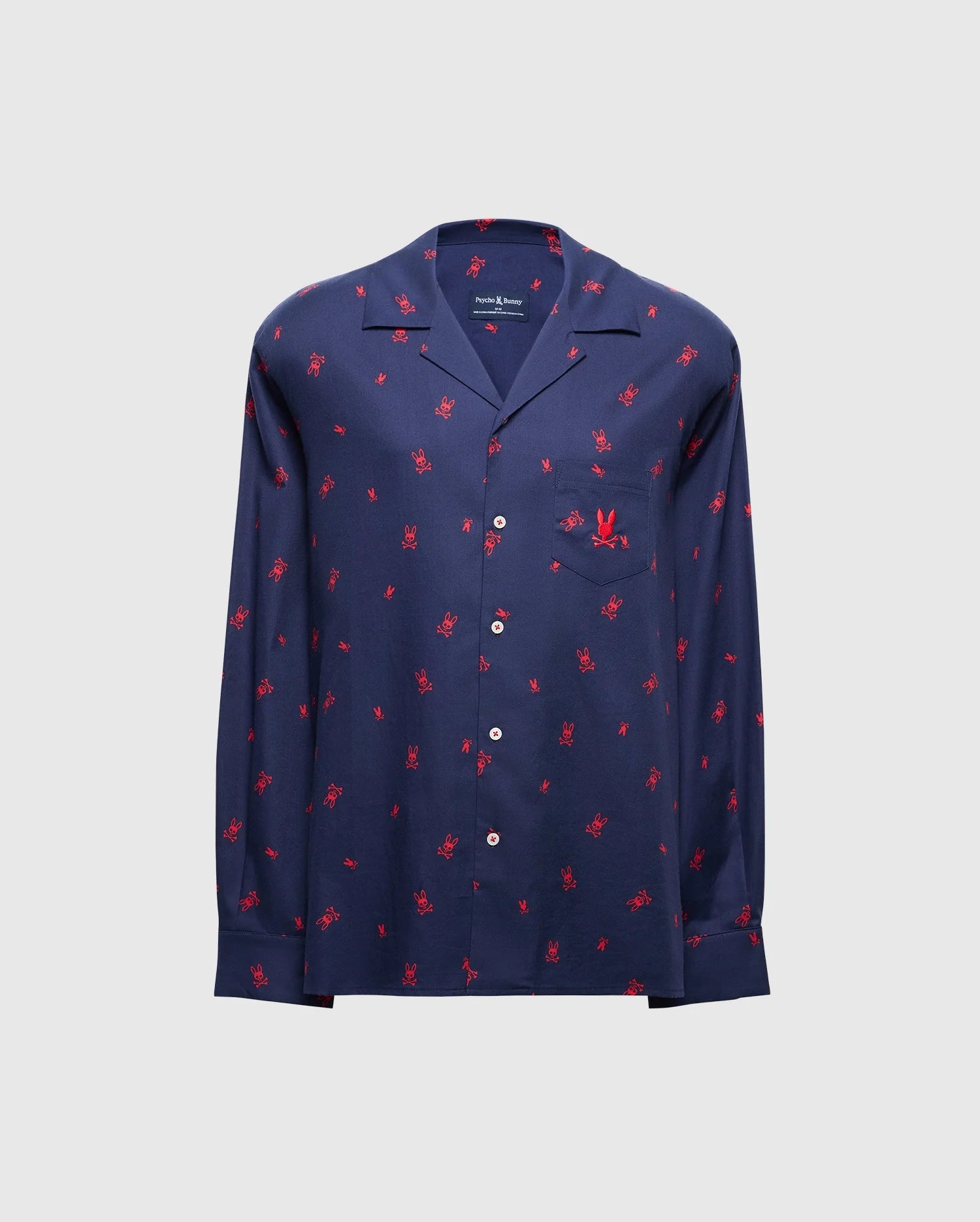 The MENS ALL OVER PRINT FLANNEL PAJAMA SET - B6X343E200 from Psycho Bunny is a stylish navy blue button-up shirt adorned with a vibrant red palm tree print on a light gray background. Designed for maximum comfort, it resembles a soft flannel sleep set and features long sleeves and a chest pocket.