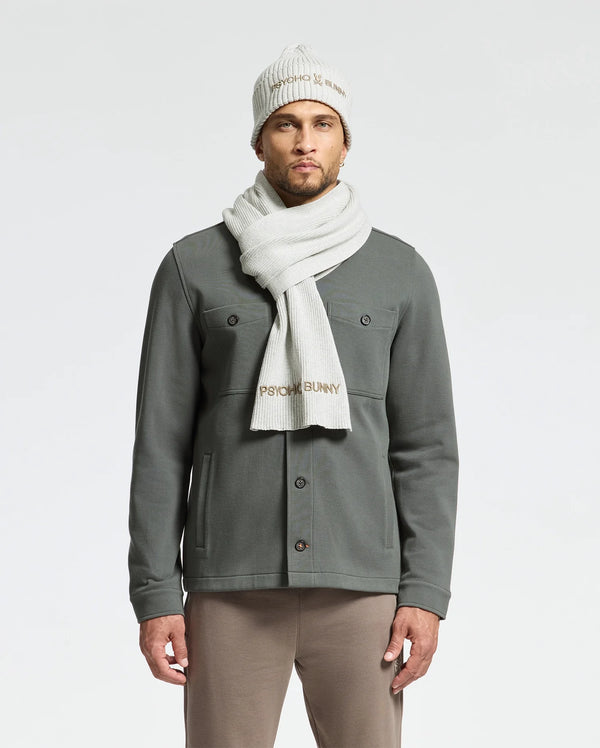 A person dons the MENS BEANIE AND SCARF BOX SET - B6X353E200 by Psycho Bunny, standing against a plain backdrop. Made from premium 100% cotton, they're clad in a gray button-up shirt and beige pants, radiating effortless style.