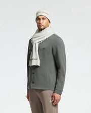 A man in a gray jacket and beige pants stands against a light background, wearing the stylish Psycho Bunny Men's Beanie and Scarf Box Set - B6X353E200. This cozy cotton set keeps him warm as he gazes forward with one hand casually tucked in his pocket.