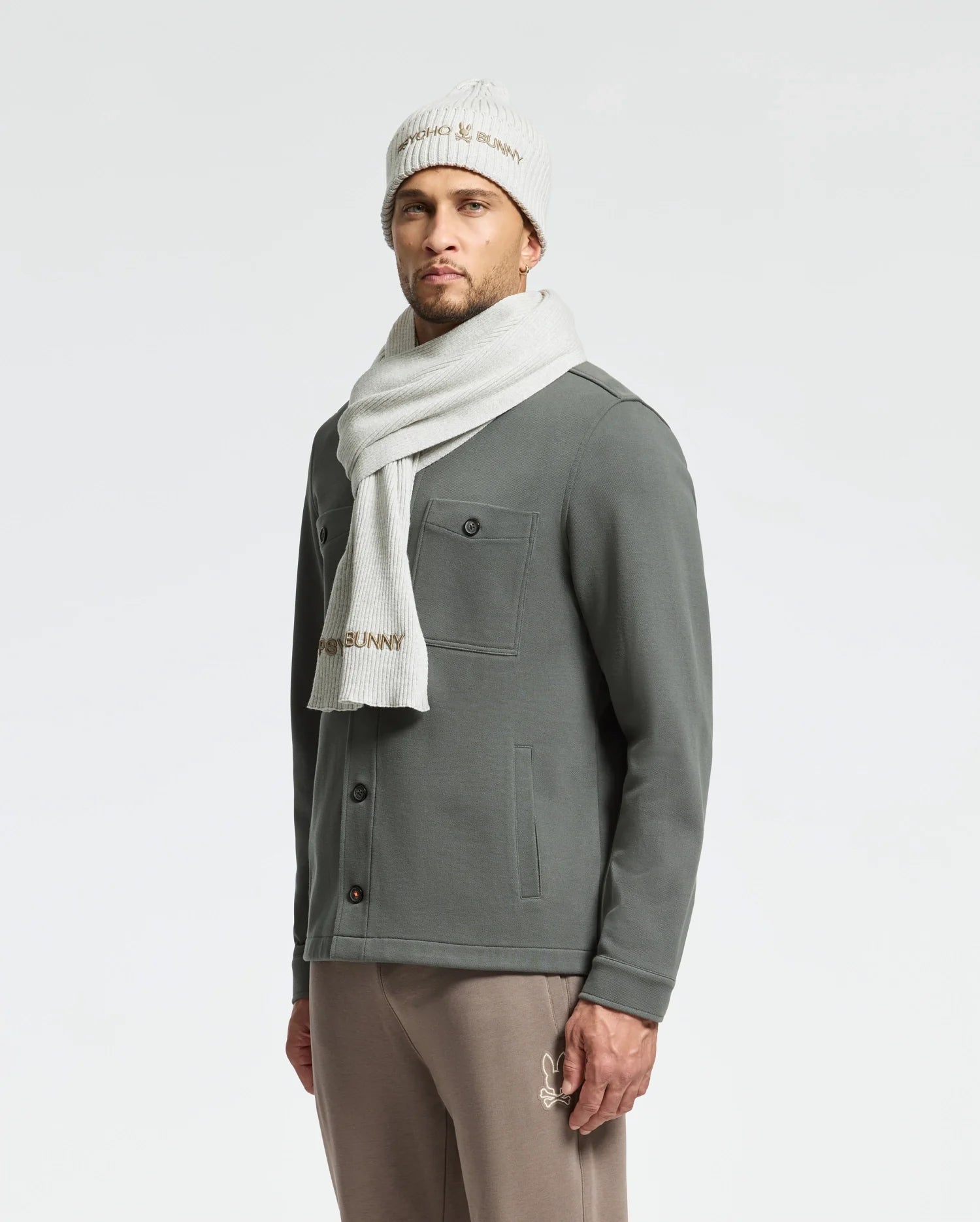 A man in a gray jacket and beige pants stands against a light background, wearing the stylish Psycho Bunny Men's Beanie and Scarf Box Set - B6X353E200. This cozy cotton set keeps him warm as he gazes forward with one hand casually tucked in his pocket.