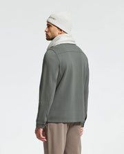 A man stands with his back turned, adorned in a Psycho Bunny MENS BEANIE AND SCARF BOX SET - B6X353E200 crafted from premium 100% cotton. The cozy set, in light gray, pairs seamlessly with his dark gray jacket and beige pants. His hands relax by his sides against a simple, light background, highlighting the luxurious comfort of this ensemble.