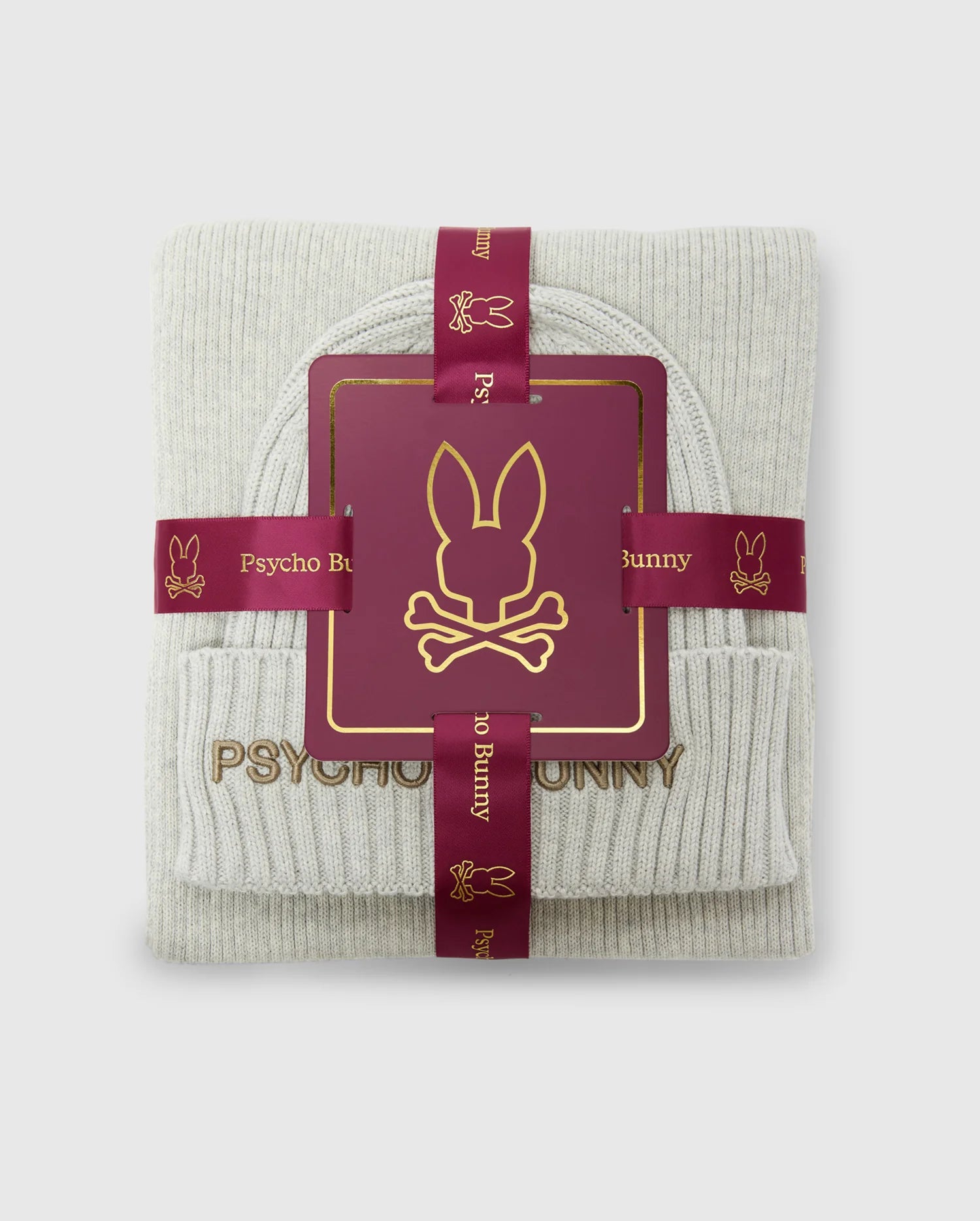 Introducing the MENS BEANIE AND SCARF BOX SET - B6X353E200 by Psycho Bunny, featuring a sleek folded grey design. This elegant gift set is wrapped in a burgundy ribbon adorned with the iconic Psycho Bunny logo. Crafted from premium 100% cotton for exceptional comfort, the set includes a logo card and is presented against a light background.