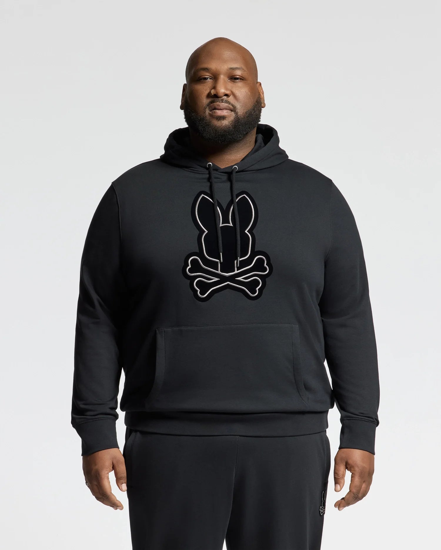 A bearded man confidently stands against a plain background, wearing the Psycho Bunny BIG AND TALL ALEXANDER HOODIE - B9H154E200. This eye-catching black pullover hoodie features a striking bunny and crossbones design on the front. Complemented by matching black pants, it offers a stylish choice perfect for big and tall individuals.