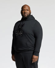 A bearded man stands against a plain backdrop wearing the comfy BIG AND TALL ALEXANDER HOODIE - B9H154E200 by Psycho Bunny, featuring a black pullover design embellished with a bunny and crossed bones motif, and looks at the camera expressionlessly.